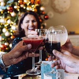 Four hands raise various cocktails in a toast, with a festive Christmas tree adorned with lights and ornaments in the background.