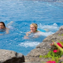 Two ladies swimming in a bright blue pool surrounded by lush greenery and vibrant flowers, creating a serene and relaxing atmosphere.