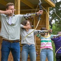 Four individuals practice archery outdoors, poised with bows drawn, focused on their targets near a wooden structure.