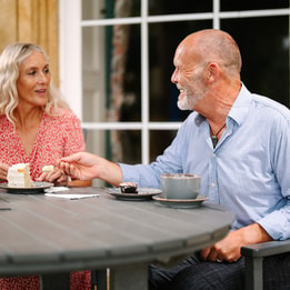 A couple enjoy dessert and coffee at a wooden table outdoors, with a cozy atmosphere and a scenic backdrop.