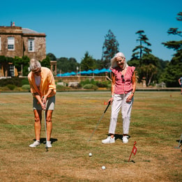 Three seniors play golf on a sunlit lawn near a historic building, focused on their shots with flags marking the holes.