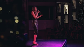 Performer singing on stage under colourful lighting in front of a seated audience.