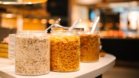 Glass jars filled with cereals and granola on a buffet counter, each with serving spoons, set against a softly lit dining area.