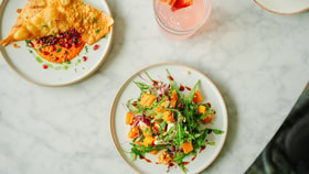 Overhead view of a salad and flatbread with dip topped with pomegranate seeds, served with a pink drink garnished with strawberries on a marble table.