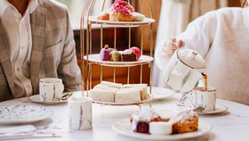 Tea being poured from a teapot into a teacup next to a tiered stand of sandwiches, cakes, and macarons, with plates of scones and jam arranged on the table.