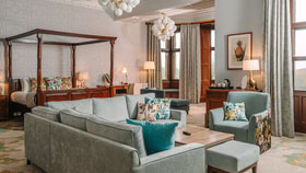 A large bedroom with neutral and pale blue decor, a dark wood four poster bed and plenty of natural light flowing through the window
