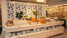 Well-lit buffet counter with plates, glasses, fruit bowls, and serving dishes, set against patterned wallpaper and a stone wall.