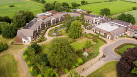 Aerial view of Alvaston Hall hotel  - a  landscaped resort with buildings arranged around gardens, pathways, and a pond, surrounded by lush green fields.