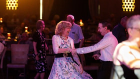 A woman in a floral dress dances with a partner in the warmly lit entertainment lounge at Alvaston Hotel, filled with seated guests.