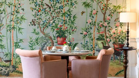 Stylish dining area with a round table and upholstered chairs set against a decorative mural of flowering trees, birds and bamboo.