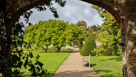 View through a brick archway onto a landscaped garden with a gravel pathway, wooden bench, manicured lawns and mature trees under a cloudy sky.