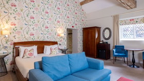 Spacious hotel bedroom with floral wallpaper, exposed wooden beam, king-size bed, blue sofa and seating area beside a leaded-glass window.