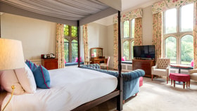 Elegant hotel suite featuring a four-poster bed, large leaded windows, comfortable lounge seating and garden views in a bright, historic setting.