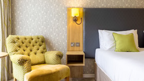 Elegant hotel bedroom featuring a tufted yellow armchair, bedside table and lamp, with a neatly made bed and patterned wallpaper.