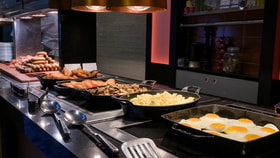Hot breakfast buffet with trays of fried eggs, scrambled eggs, mushrooms, tomatoes, sausages, and hash browns under heat lamps.