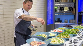 Chef in a kitchen uniform sprinkling herbs over large bowls of fresh salads at a buffet counter.