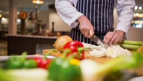 Chef slicing an onion on a wooden chopping board, surrounded by fresh vegetables including bell peppers and leeks on a kitchen counter.