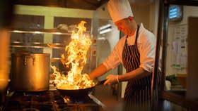 Chef in a professional kitchen cooking with a pan over open flames on a gas stove, with a large pot beside it and industrial kitchen equipment in the background.