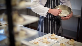 Chef whisking a mixture in a metal bowl while plating small desserts drizzled with sauce on rectangular plates.