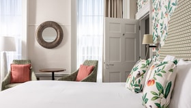 A bright bedroom seating area with two upholstered chairs and a small round table beside a window, next to a neatly made bed with botanical cushions and patterned wallpaper.