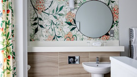 A bathroom with a round mirror above a wide sink shelf, wood-toned tiles, and floral-patterned wallpaper, with neatly folded towels and toiletries.