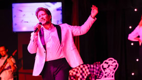 Performer in a light blazer singing into a microphone under pink stage lighting.