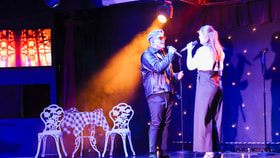 Two singers face each other on stage with microphones, lit by bright coloured lights.