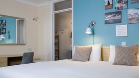 Bright hotel room with a white bed and textured cushions, a blue feature wall with coastal artwork, a wall-mounted lamp, desk with mirror, and an open doorway to an ensuite bathroom.