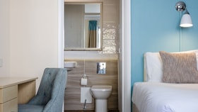 Hotel room with a white bed beside a blue accent wall, next to an open ensuite bathroom featuring a toilet, sink, and mirror, with a desk and upholstered chair in the foreground.