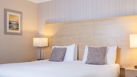 Modern hotel bedroom with a large white bed, wooden headboard, textured cushions, soft bedside lighting, and neutral-toned walls with framed artwork.