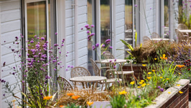 Outdoor garden seating area with round tables and chairs set among colourful flowers and plants, alongside a row of light blue lodge-style buildings with glass doors.