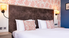 Modern bedroom with a large white bed, coral-patterned accent cushions, a dark headboard, and warm bedside lighting set against a bold patterned feature wall.