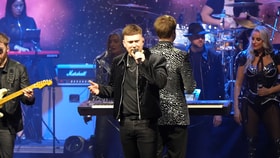 Live stage performance featuring singers, musicians and dancers under colourful concert lighting, with keyboards, guitar and drum kit visible.