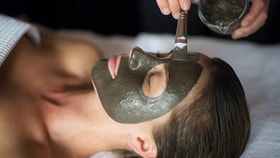 A woman lies relaxed with a grey facial mask applied, while a therapist brushes on more product in a spa setting.