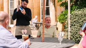 A waiter in a black shirt shakes a cocktail shaker at an outdoor table, while guests enjoy drinks under a sunny sky a Cricket St. Thomas