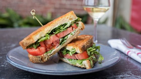 A close-up of a grilled sandwich with fresh tomatoes, greens, and a glass of white wine in the background, served on a gray plate.