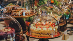 Display of cakes and pastries arranged on wooden stands and covered with glass domes, including a sponge cake decorated with strawberries and cream, pastries drizzled with icing, and other desserts set against a softly lit café background with greenery and blurred lights.