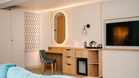 Contemporary hotel room featuring a wooden desk and dressing area with an illuminated mirror, integrated minibar and tea-making facilities. A wall-mounted television, patterned feature wallpaper and soft lighting create a modern, comfortable atmosphere.
