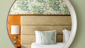 A neatly made bed with white linens and striped cushions, a padded beige headboard, and a wooden bedside panel with a wall lamp, framed through a round mirror against leafy patterned wallpaper.