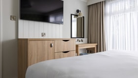 A tidy bedroom corner with a wall-mounted TV above light wood cabinetry, next to a small desk with a mirror and stool, set by a window with sheer curtains.