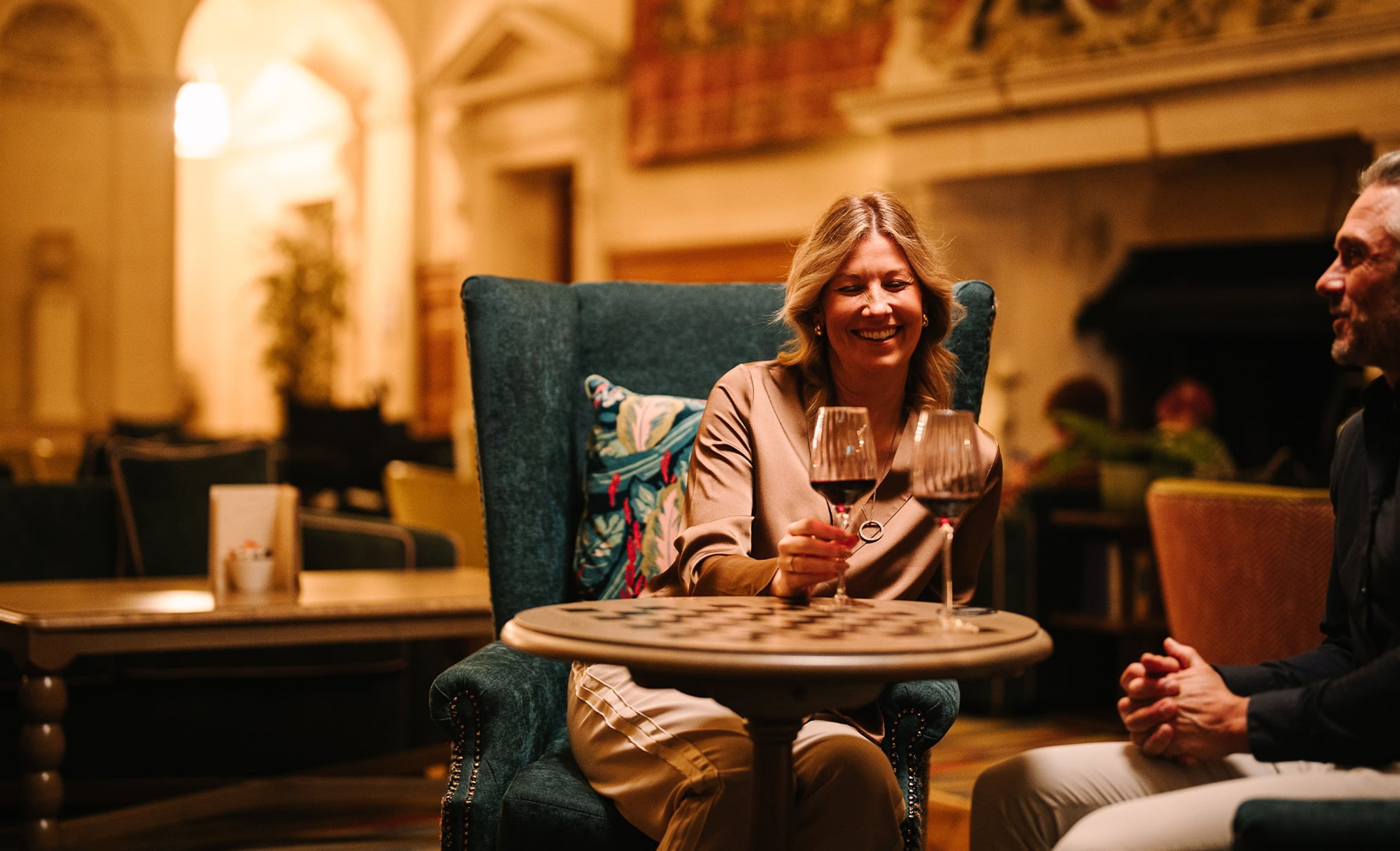 A warmly lit lounge setting with two people seated at a small round table, each holding a glass of red wine. The space features upholstered chairs, patterned cushions, and soft ambient lighting, with blurred seating and architectural details in the background creating a cosy atmosphere.