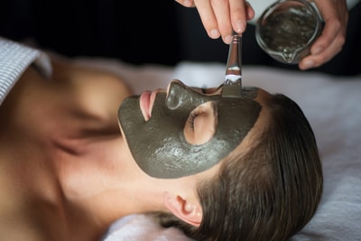 A woman lies relaxed with a grey facial mask applied, while a therapist brushes on more product in a spa setting.
