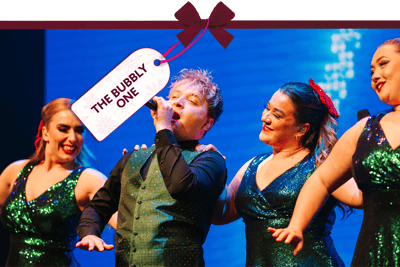 Festive stage performance featuring a singer holding a microphone, surrounded by performers in sequinned outfits under bright blue lighting. A gift tag tied with a red ribbon reads ‘The Bubbly One’.