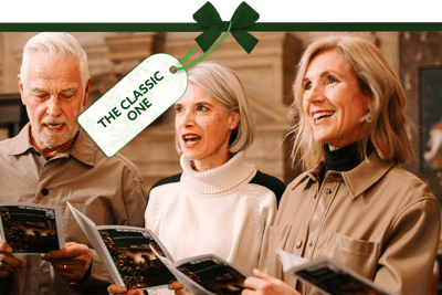 Festive indoor scene with three people holding songbooks and singing together in a traditional setting. A gift tag tied with a green ribbon reads ‘The Classic One’