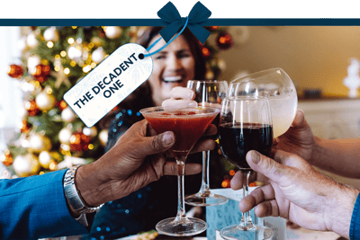 Festive scene with hands raising cocktails and wine glasses in a toast in front of a decorated Christmas tree, with a blurred figure in the background. A gift tag tied with a blue ribbon reads ‘The Decadent One’