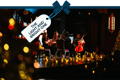 Candlelit performance with a small string ensemble seated on stage, surrounded by glowing candles and warm lights. A gift tag tied with a blue ribbon reads ‘The Light and Bright One’