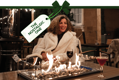 Festive outdoor setting with a person seated beside a fire pit, wearing a cosy coat, with drinks on the table and warm lighting in the background. A gift tag tied with a green ribbon reads ‘The Nostalgic One’