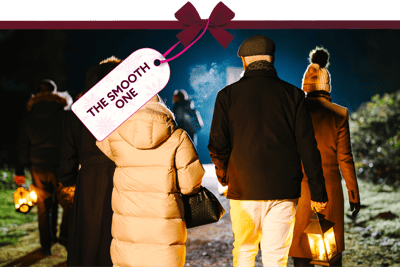 Evening outdoor scene showing people walking along a lit path holding lanterns, dressed in warm winter clothing. A gift tag tied with a red ribbon reads ‘The Smooth One’