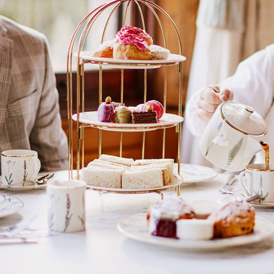 Tea being poured from a teapot into a teacup next to a tiered stand of sandwiches, cakes, and macarons, with plates of scones and jam arranged on the table.