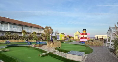 A colorful mini-golf course featuring a striped lighthouse, beach huts, and lush greenery, with playful blue and green pathways.